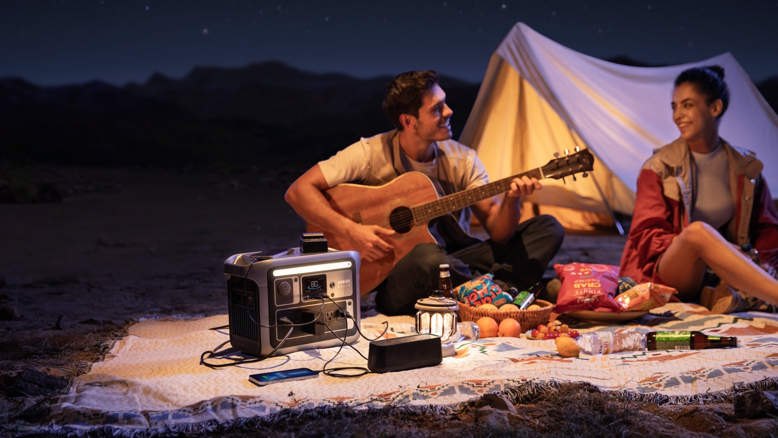 Two campers with a guitar beside a lit tent at night